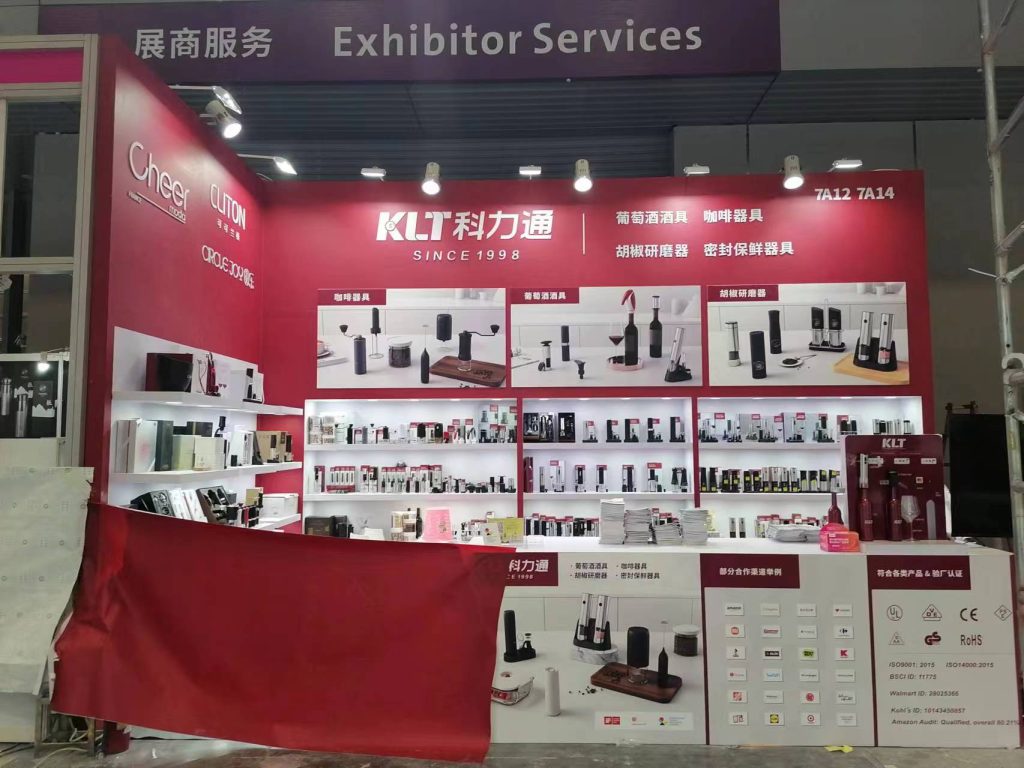 Red and white exhibition booth displaying kitchen gadgets on shelves. A counter shows various products and certifications. Red fabric is draped below.