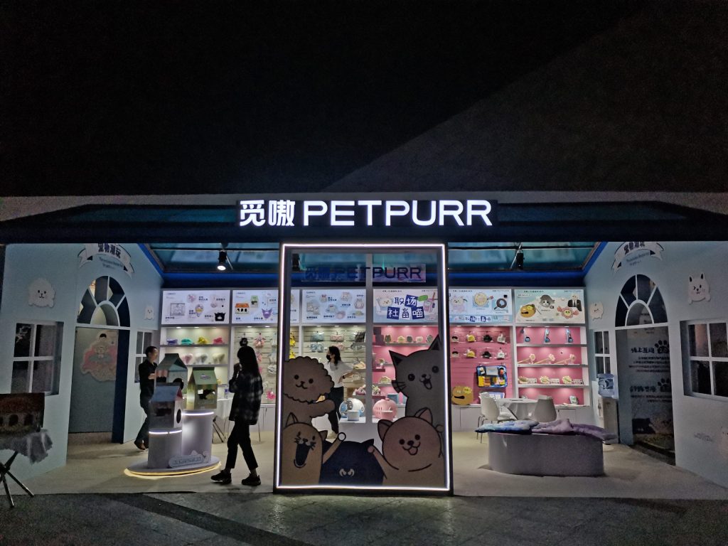 Front view of a brightly lit house-shaped booth at night, filled with various products and featuring large cartoon animal faces. People are inside.