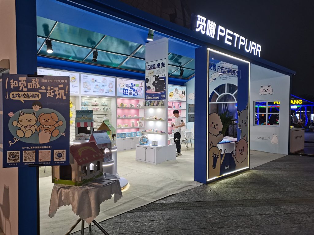 View into a brightly lit house-shaped booth with shelves of products, cartoon animal displays, and small decorative house models on a table.