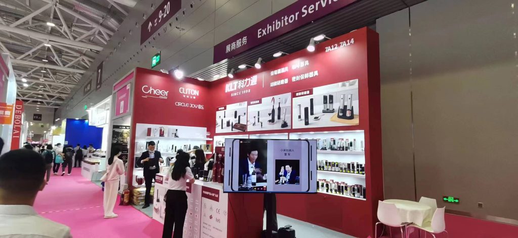 Red and white exhibition booth with displayed kitchen gadgets. Visitors are walking by, and a large phone screen on a stand shows a video.