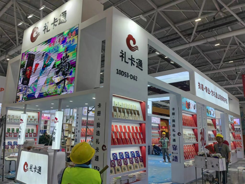 A large exhibition booth with a glitching digital screen displaying abstract art, surrounded by shelves of products. Workers in yellow hard hats are present.