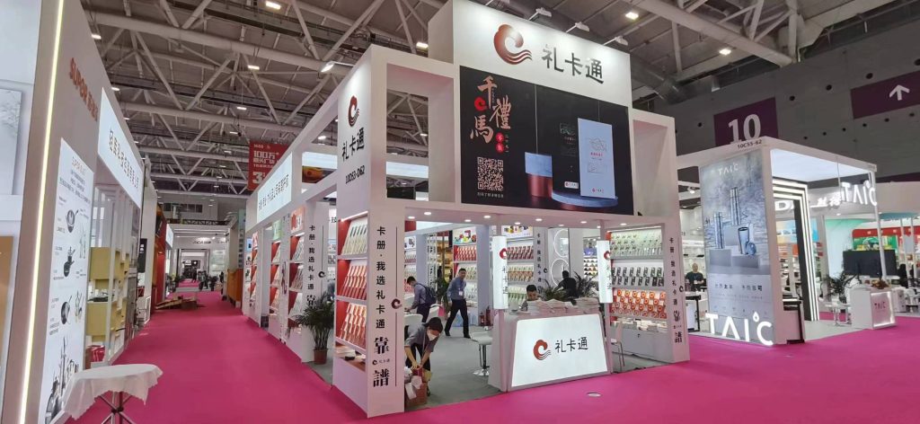 A large white exhibition booth with product displays and a screen showing merchandise in a busy convention hall with pink carpet.
