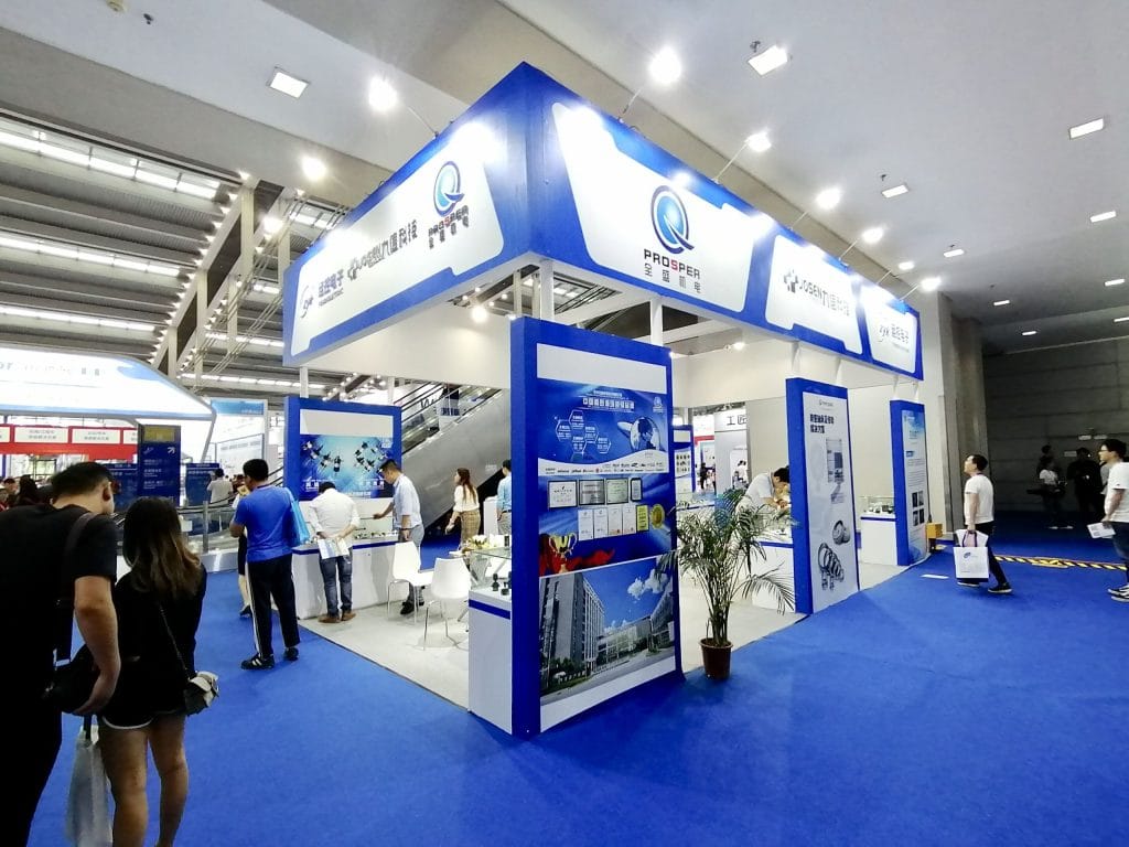 Bustling trade show floor with many people around a blue and white exhibition booth displaying electronic components. Blue carpet covers the floor.