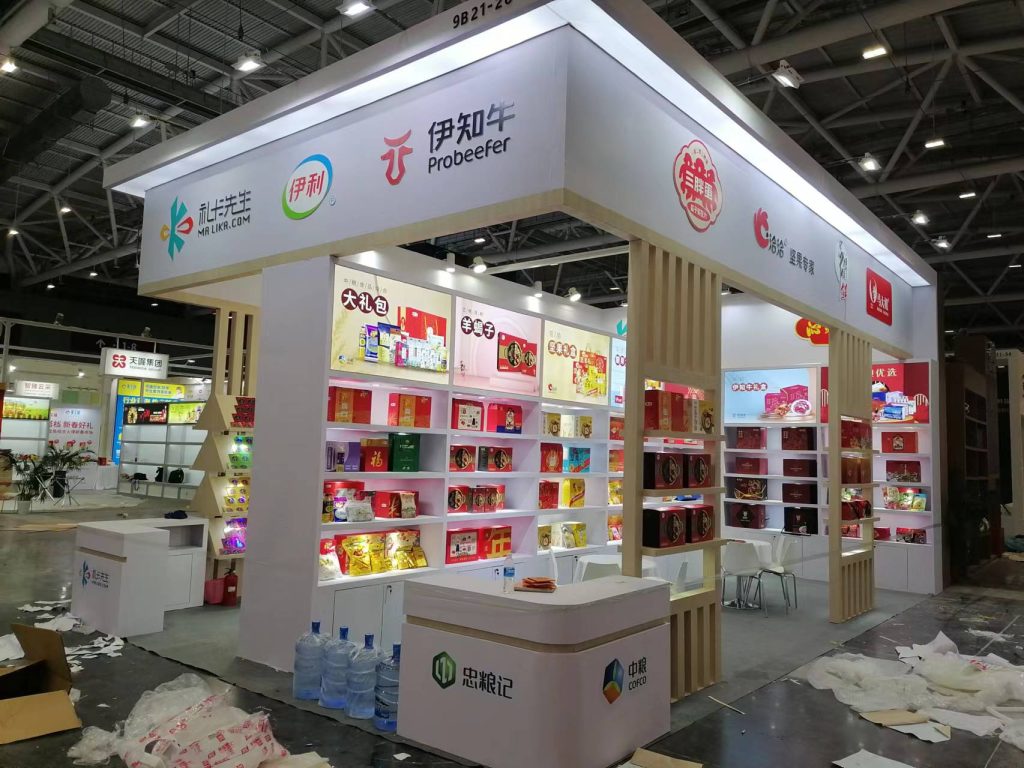 A white and wood trade show booth with shelves displaying numerous packaged food products. Construction debris litters the unfinished floor.