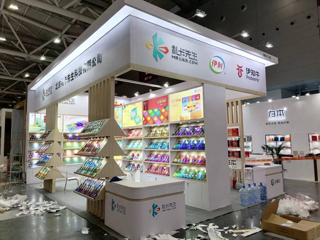 A brightly lit trade show booth showcasing shelves and a triangular display with colorful, reflective packaged products. Debris is on the unfinished floor.