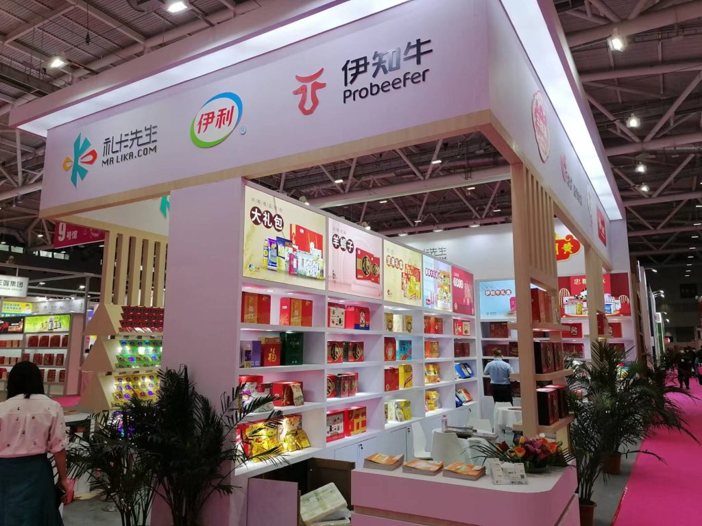 A trade show booth with shelves full of packaged food products, many in red and gold boxes. People walk on a pink carpeted aisle past potted plants.