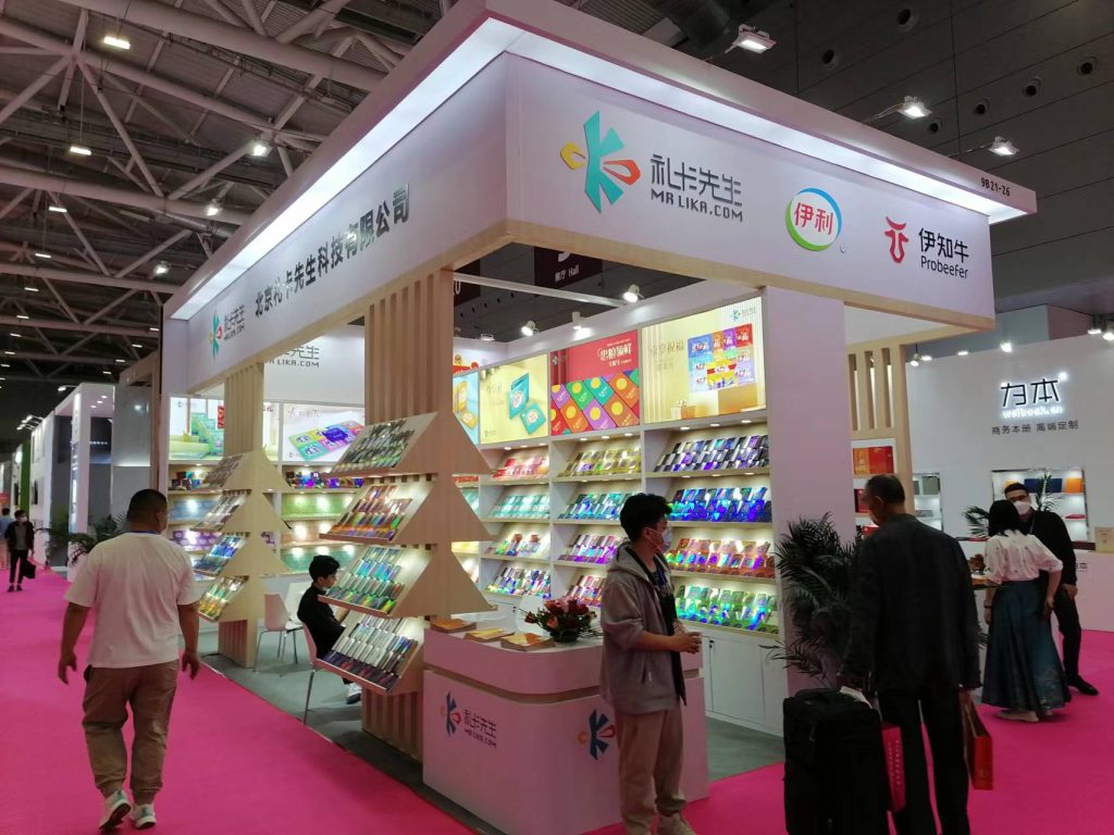 A brightly lit trade show booth displays colorful packaged products on shelves and a triangular rack. People walk on a pink carpeted aisle.
