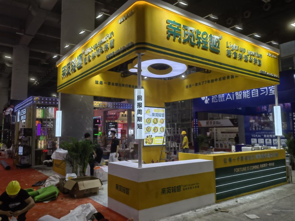 Yellow and green exhibition booth under construction, with workers in hard hats and building materials on a red floor.