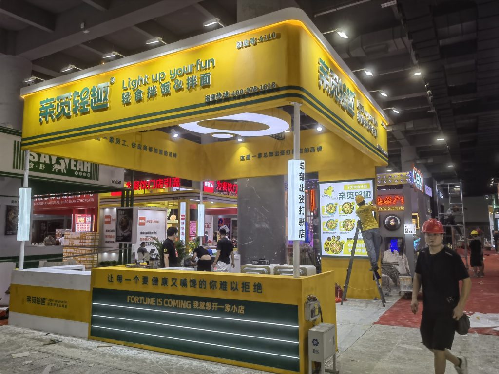 Exhibition booth in yellow and green being assembled, with workers, a menu screen displaying food, and construction materials on the ground.