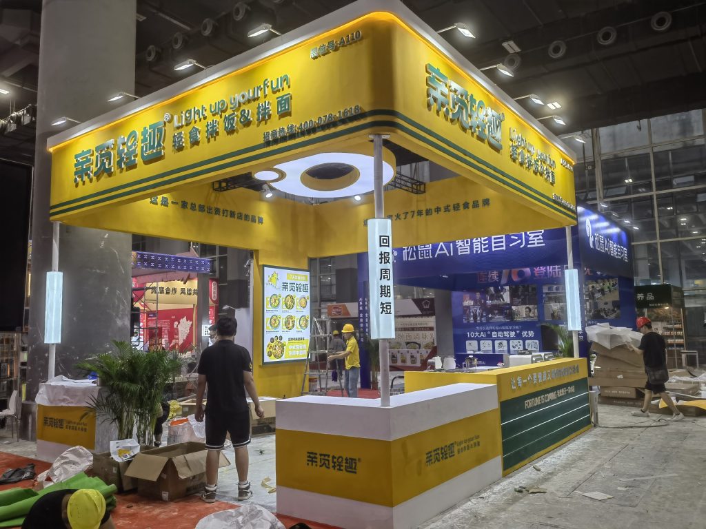 Yellow and green exhibition booth under assembly with workers, a digital menu displaying food, and scattered construction debris on the floor.