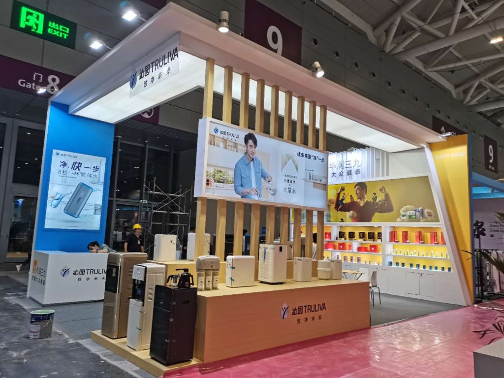 A large exhibition booth features water purifiers and appliances on wooden stands. A man on a screen, and a worker, are visible.