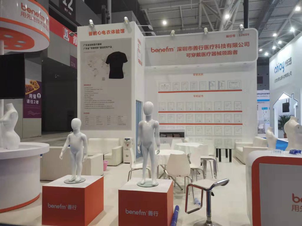 Trade show booth featuring white walls, orange accents, mannequins, a black t-shirt display, and wall-mounted certificates.