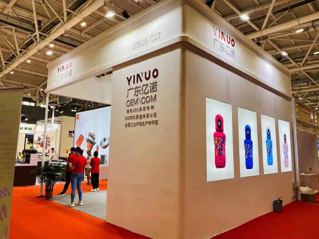 Trade show booth with white walls, displaying colorful water bottles and Chinese text. Several attendees are visible on a red carpet.