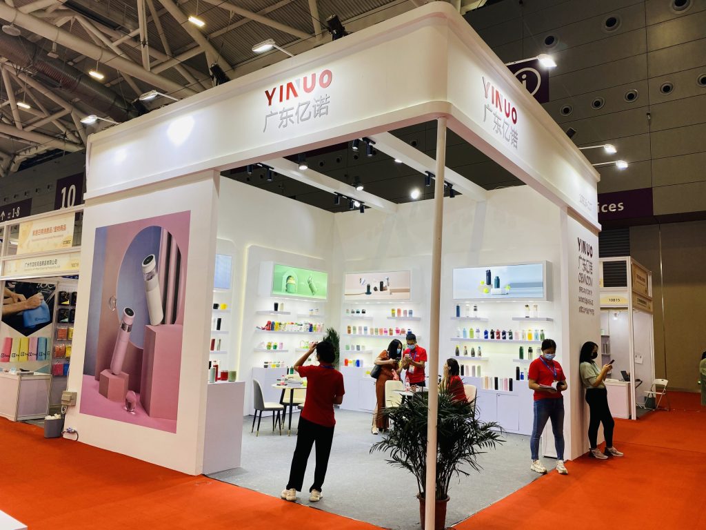 Busy trade show booth with white walls, shelves displaying various products, and several people on a red carpet.