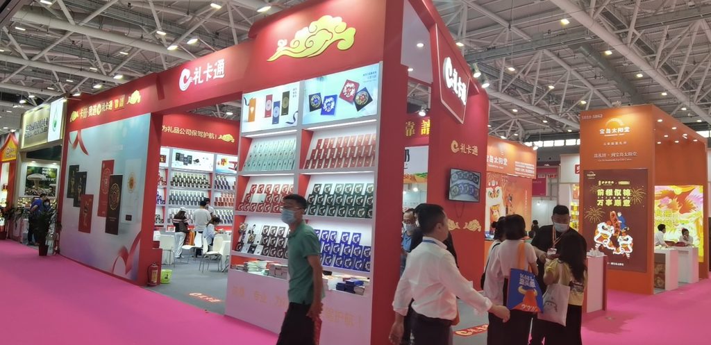 Red exhibition booth at a trade show, displaying many packaged products on shelves, with people walking on a pink carpet.
