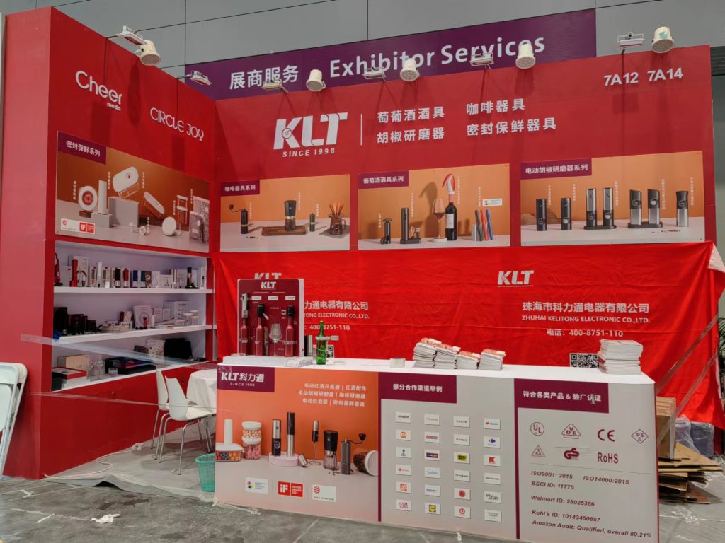 Busy red exhibition booth with display shelves full of products. People are gathered around a white counter, which also displays items.