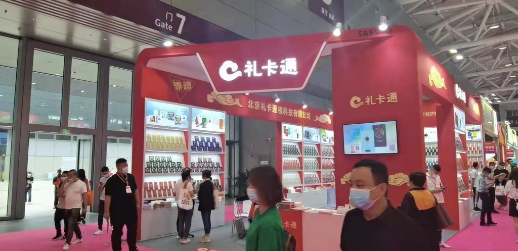 Red exhibition booth inside a large hall, showcasing products on shelves. People in face masks are moving through the bright trade show environment.