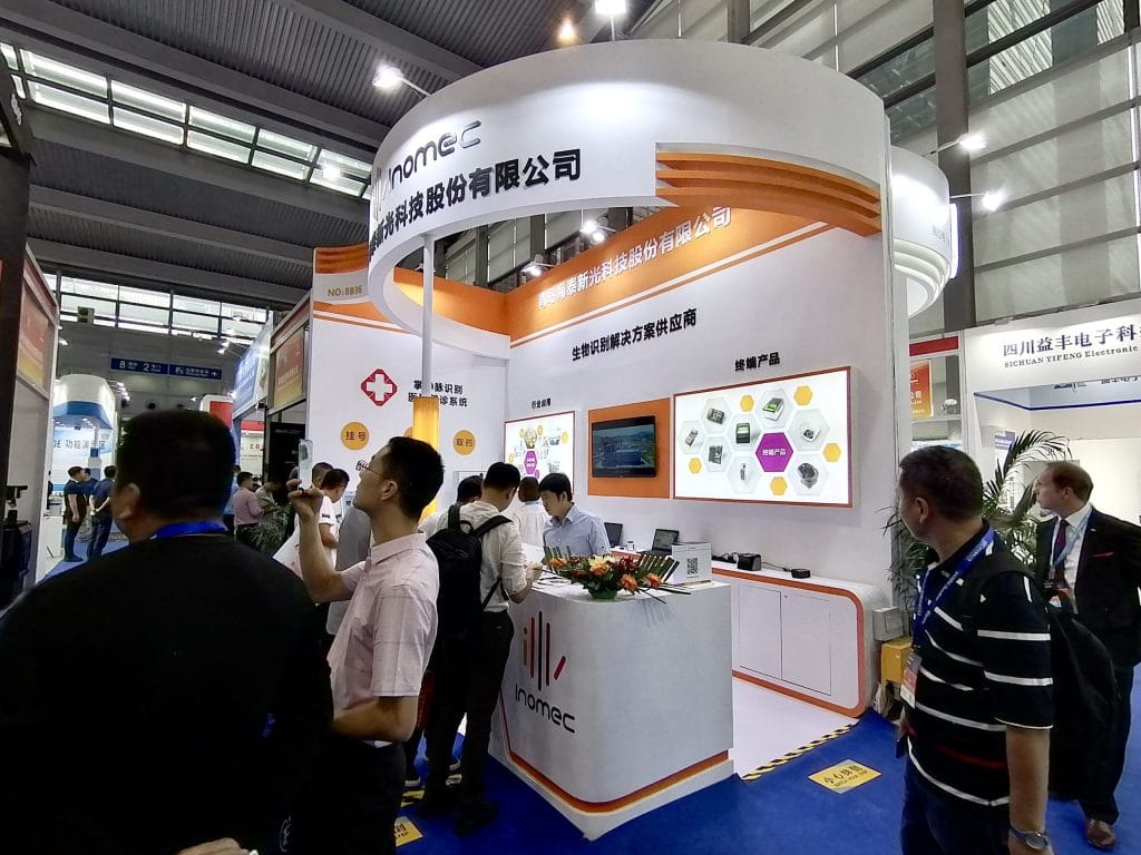 People gather at a brightly lit exhibition booth, featuring white and orange design, product displays, and a reception counter at a trade show.
