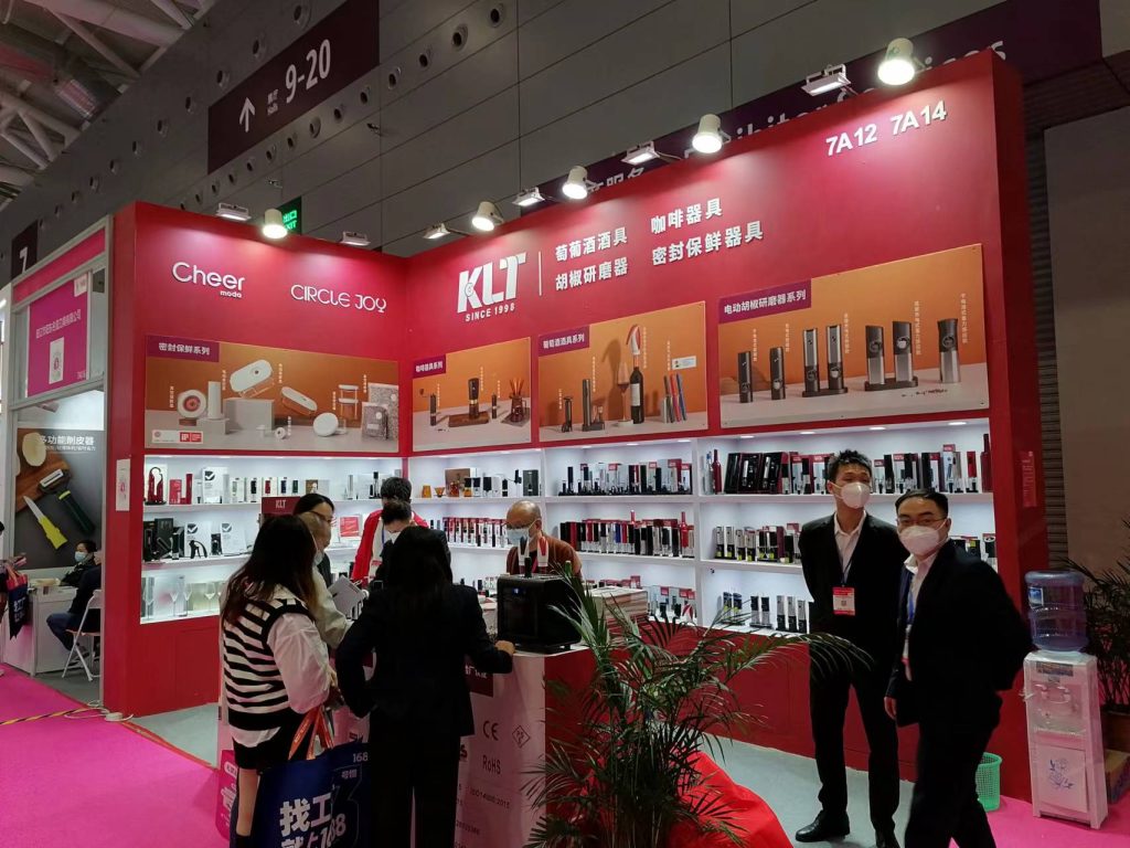 Exhibition booth with display shelves of products and multiple masked attendees. A large white counter and a plant are in the foreground.
