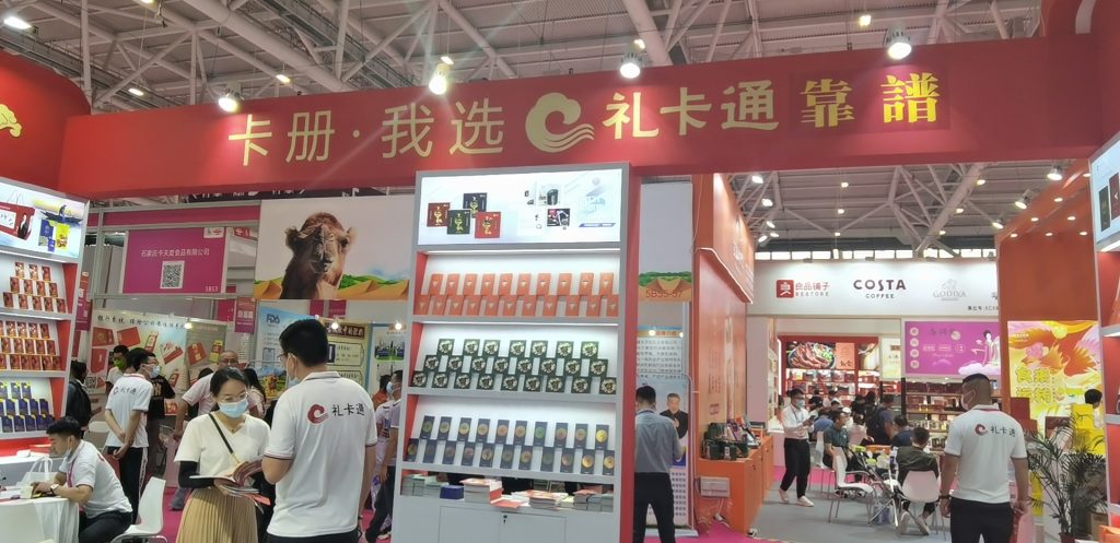 Wide view of a red exhibition booth at a trade show, full of displayed products. People are walking around the busy hall under a large red banner.