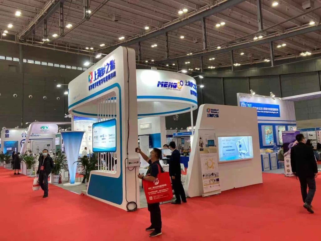 A man films a large white and blue exhibition booth with a phone at a bustling trade show.