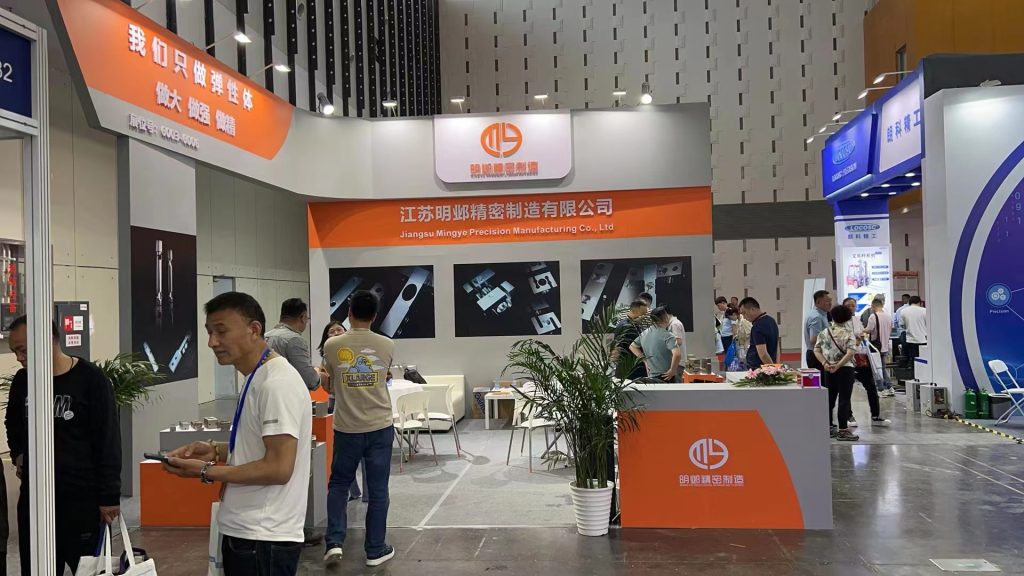 A busy trade show booth with many people. Orange and gray displays showcase industrial components on screens and tables.