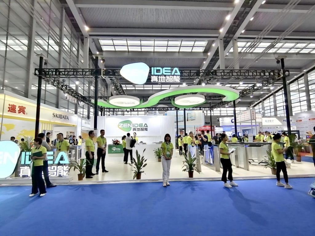 Busy trade show booth featuring a black truss structure, green curved lights, and multiple white access control devices. Many attendees and staff are present.