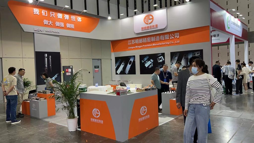 A bustling trade show booth, viewed from another angle, with visitors. Orange and gray displays feature industrial components on tables and screens.