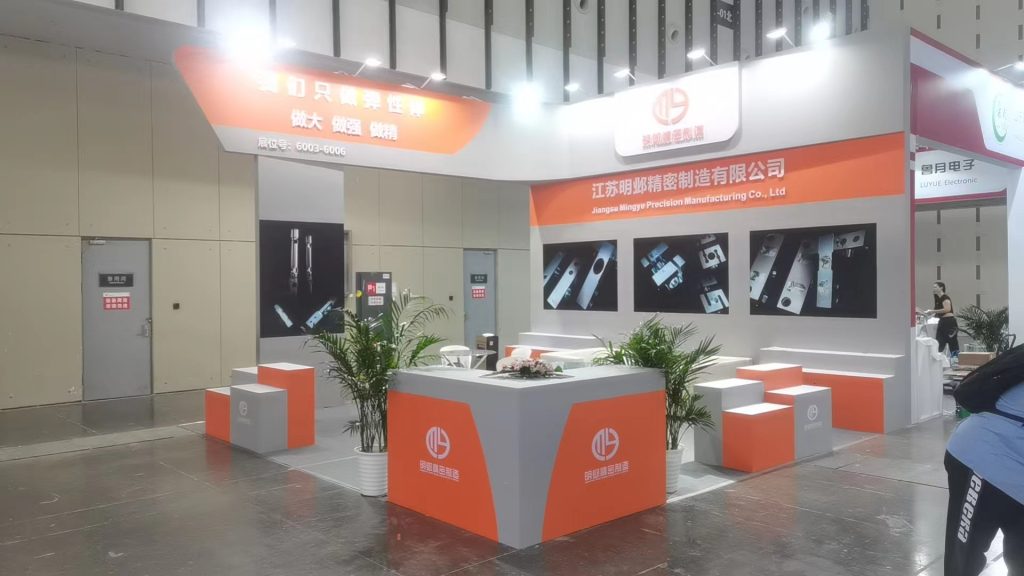 An empty trade show booth setup with orange and gray modular displays, featuring industrial components on wall screens and potted plants.