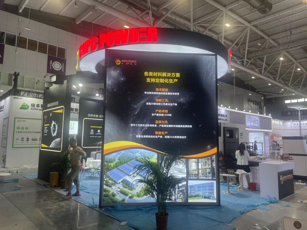 An exhibition booth in a large hall, featuring a prominent vertical display showing solar panels and industrial images. Blue protective sheeting covers the floor.