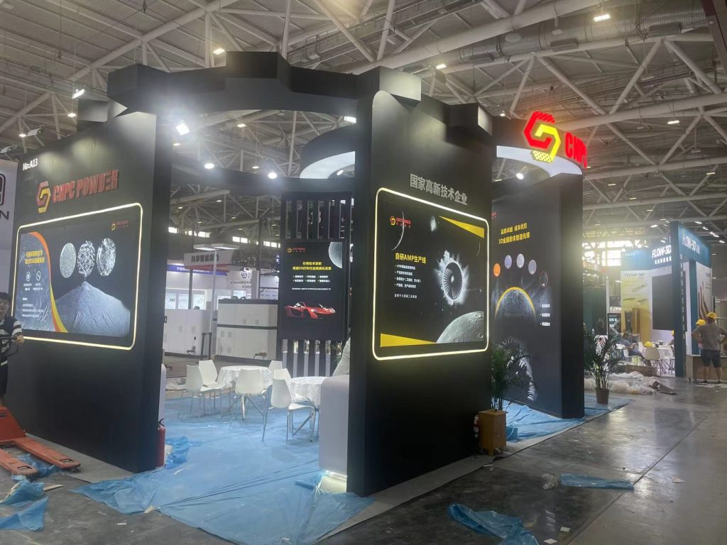 A modern exhibition booth under construction, with several illuminated screens displaying technical graphics and a seating area with white tables and chairs on a blue tarp.