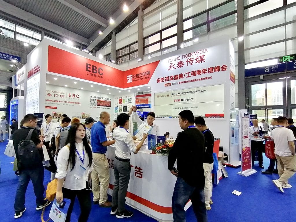 Crowded exhibition booth at a trade show, with attendees interacting around a counter. The booth features red and white branding and display screens.