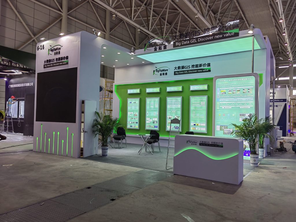 An exhibition booth under construction, with scaffolding visible and workers in the background. The white and green booth features blank screens, a reception desk, and potted plants.