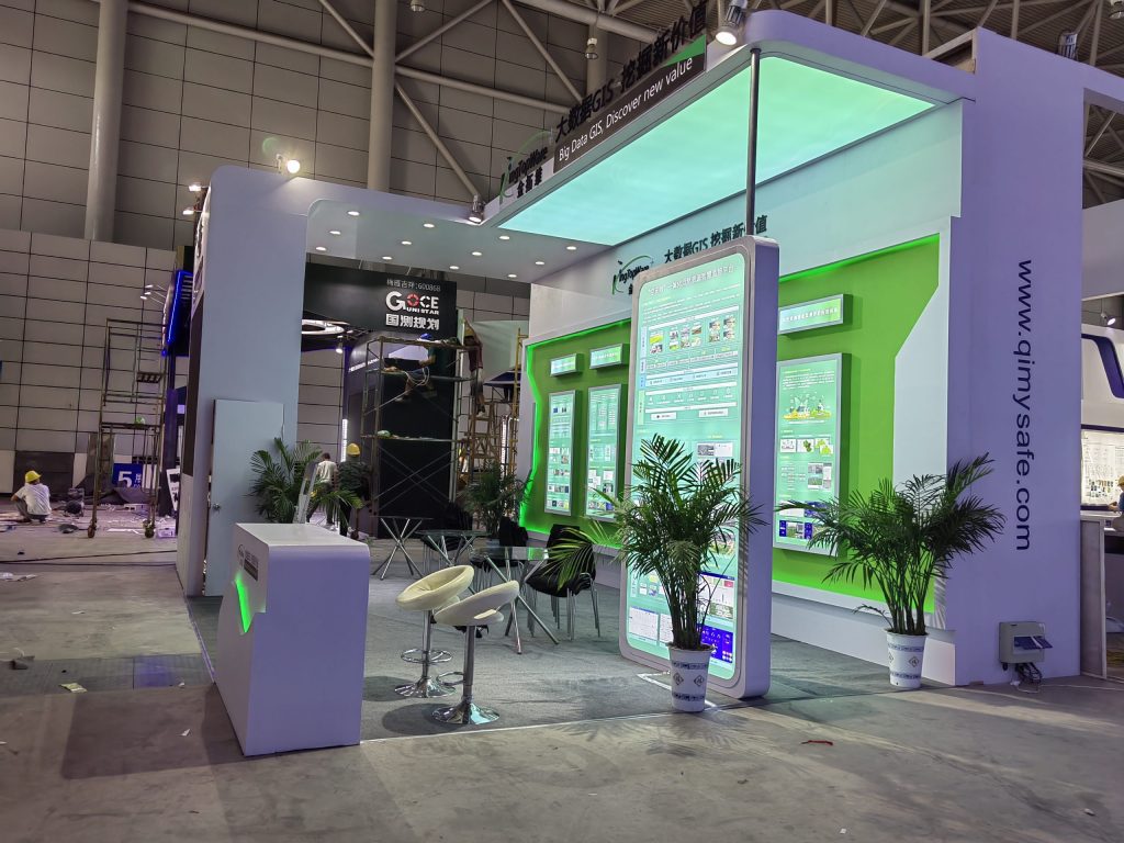 A side view of an exhibition booth under construction, showing scaffolding and workers. The white and green booth features multiple screens, some displaying content, and potted plants.