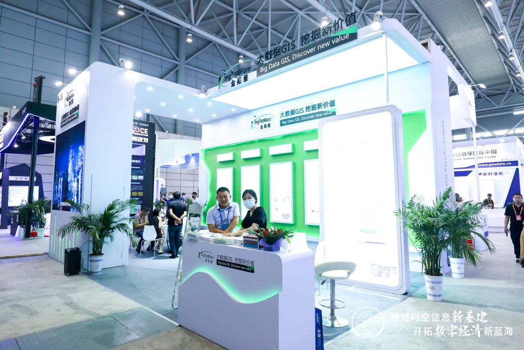 Two attendees, one masked, stand at a white reception desk within a brightly lit exhibition booth. Potted plants and display screens are visible in the green-accented space.