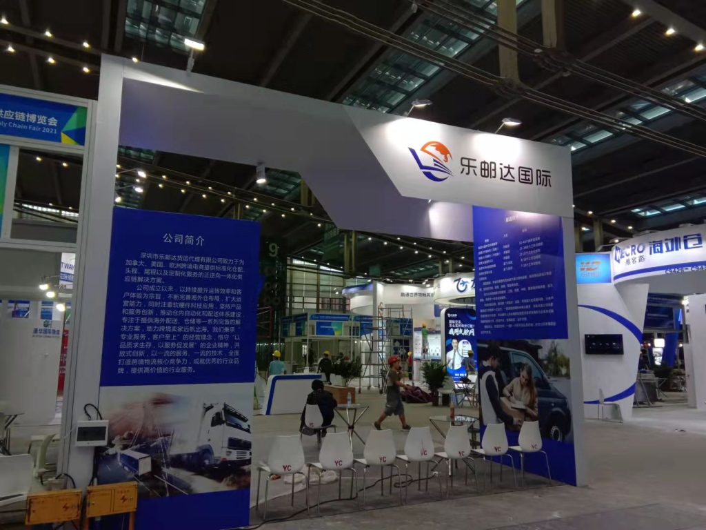 Real-world exhibition booth with blue and white design, display screens, text panels, and white chairs in a busy hall.
