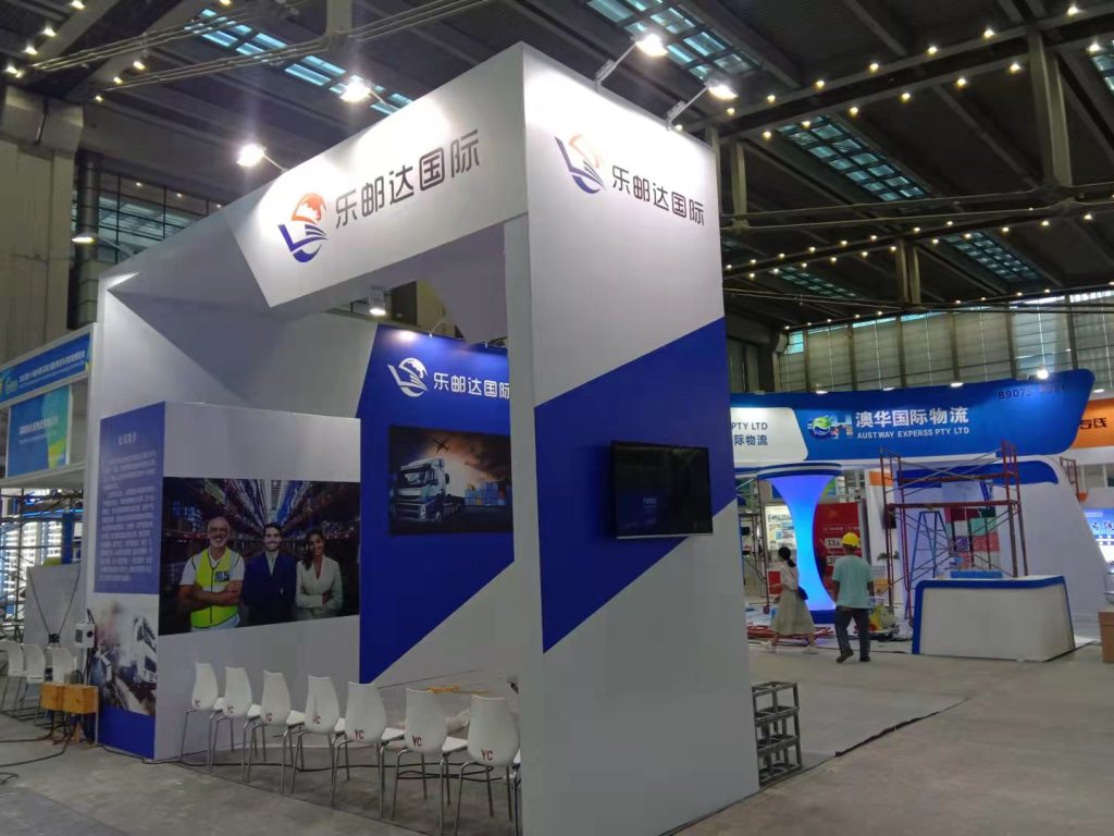 Trade show booth in blue and white, featuring screens with logistics images and smiling staff, surrounded by white chairs.