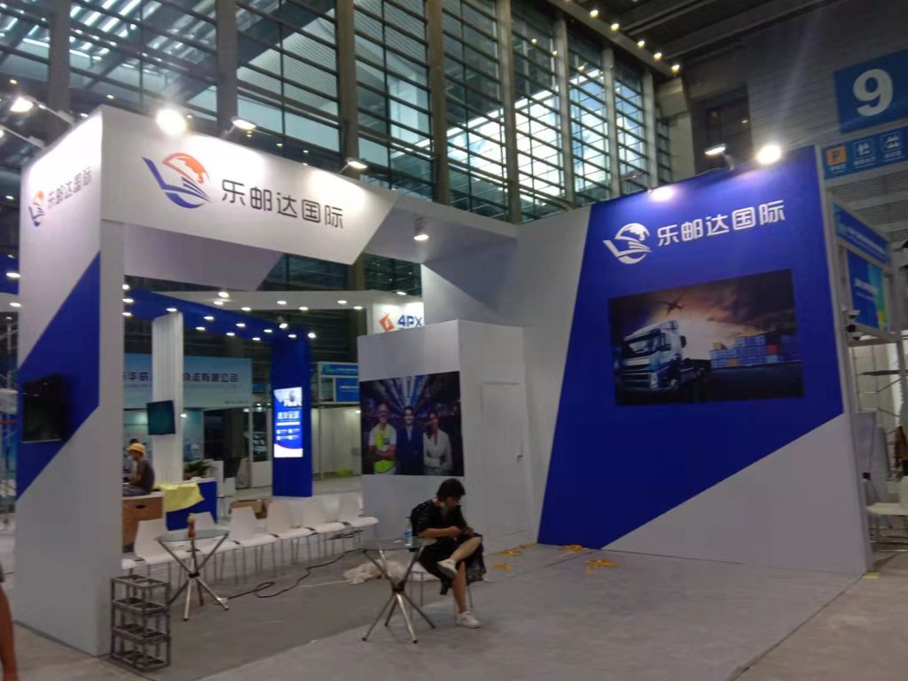 Exhibition booth with blue and white geometric design, screens showing trucks and smiling individuals. A person sits nearby.