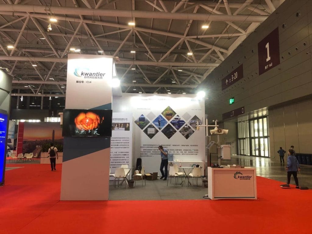 Trade show booth with a red carpet, two screens displaying a lit flame, and people walking.