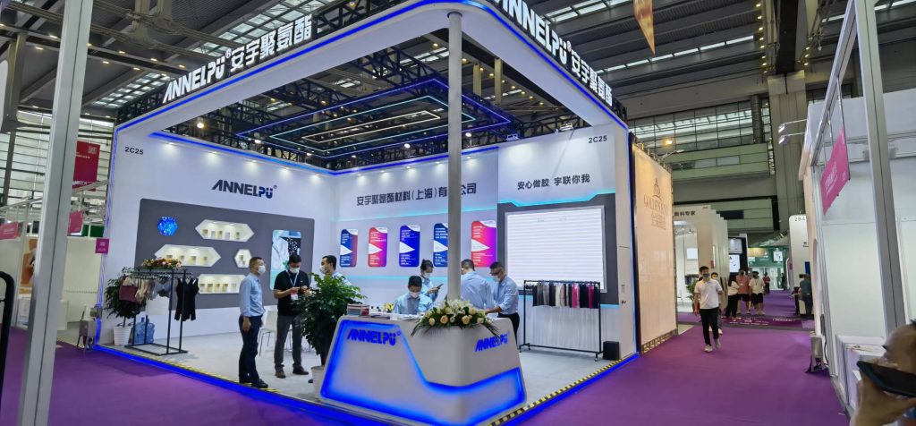 People interact at a white and blue exhibition booth displaying products and information, with several attendees and staff present.