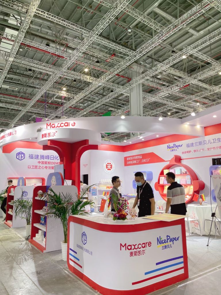 Three men in face masks interact at a red and white exhibition booth displaying numerous packaged products, under a large industrial ceiling structure.