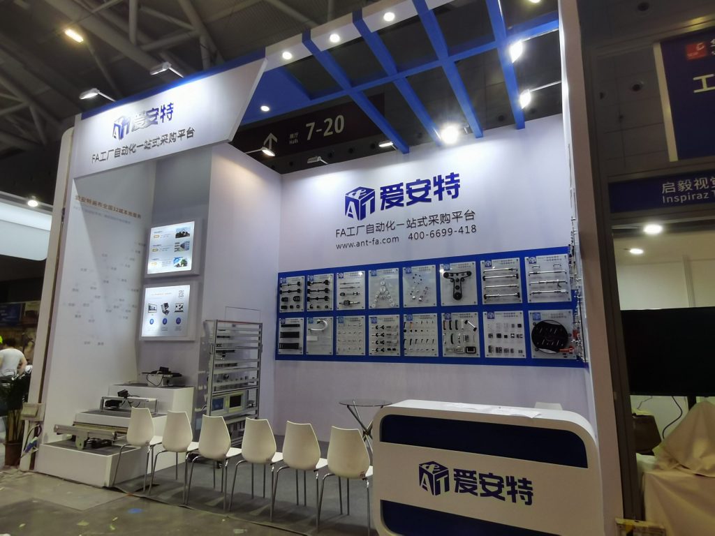 A white and blue exhibition booth featuring displays of various industrial components. A reception desk, small table with chairs, and overhead lighting are visible.