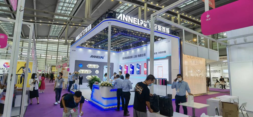 People gather at a bustling exhibition booth, interacting with staff and viewing product displays. Other booths are visible in the background.