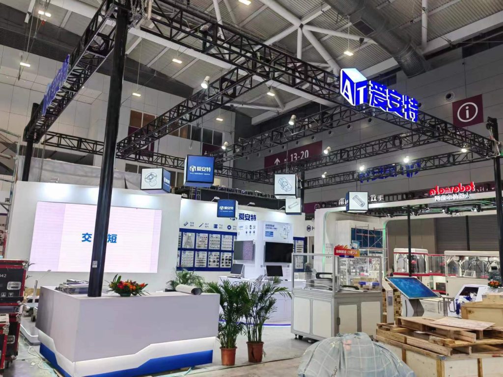 A trade show booth with white and blue accents, featuring large display screens, smaller panels with components, and a prominent overhead truss structure in a large hall.