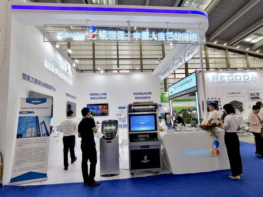 An active exhibition booth with people interacting with staff and displays. The booth is white with blue accents, featuring multiple screens and Chinese text.