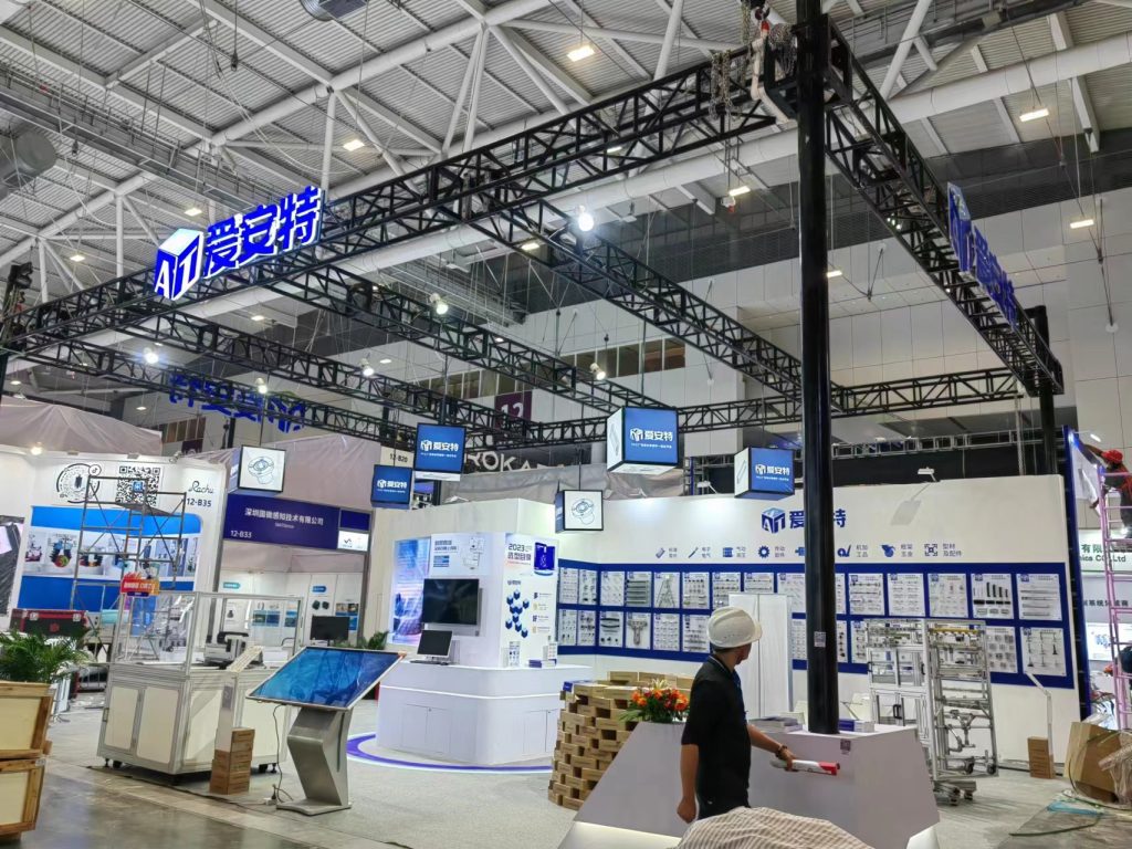 A wide view of a trade show booth with a large black truss system, various display screens and panels. A worker in a hard hat is visible at the right.