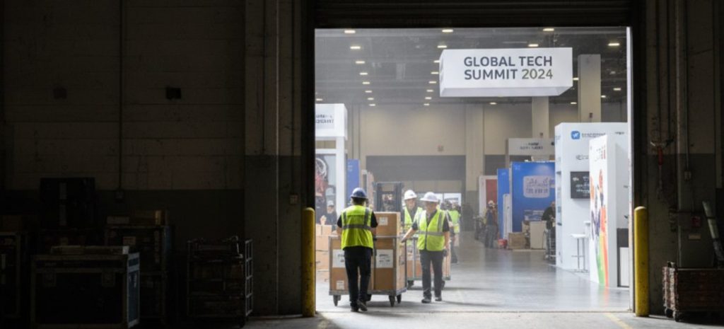 Workers moving and carrying exhibition crates and booth materials in a large convention centre, using dollies and hand trucks, wearing safety vests and gloves, realistic modern trade show environment, bright lighting, dynamic action, showing teamwork and physical effort, perspective slightly angled to capture motion and scale, high detail, professional, realistic style.