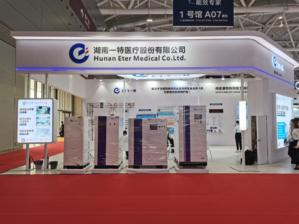 An exhibition booth with a red carpet, displaying several large white and purple medical devices wrapped in plastic, with people in the background.