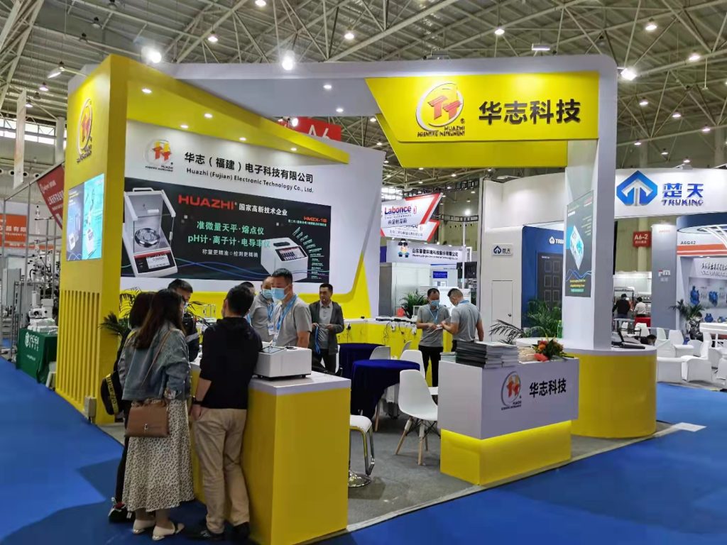 People interact at a bright yellow and white trade show booth displaying products on screens and counters in a bustling exhibition hall.