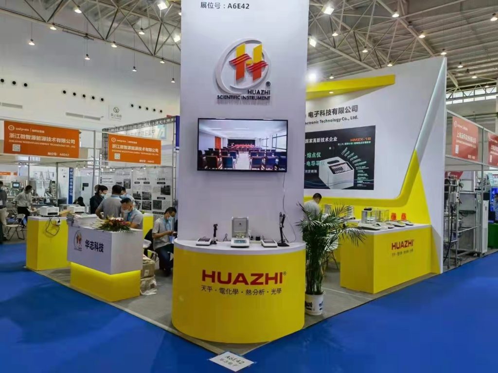 A yellow and white trade show booth with a central pillar featuring screens and product displays. Visitors are visible in the background.